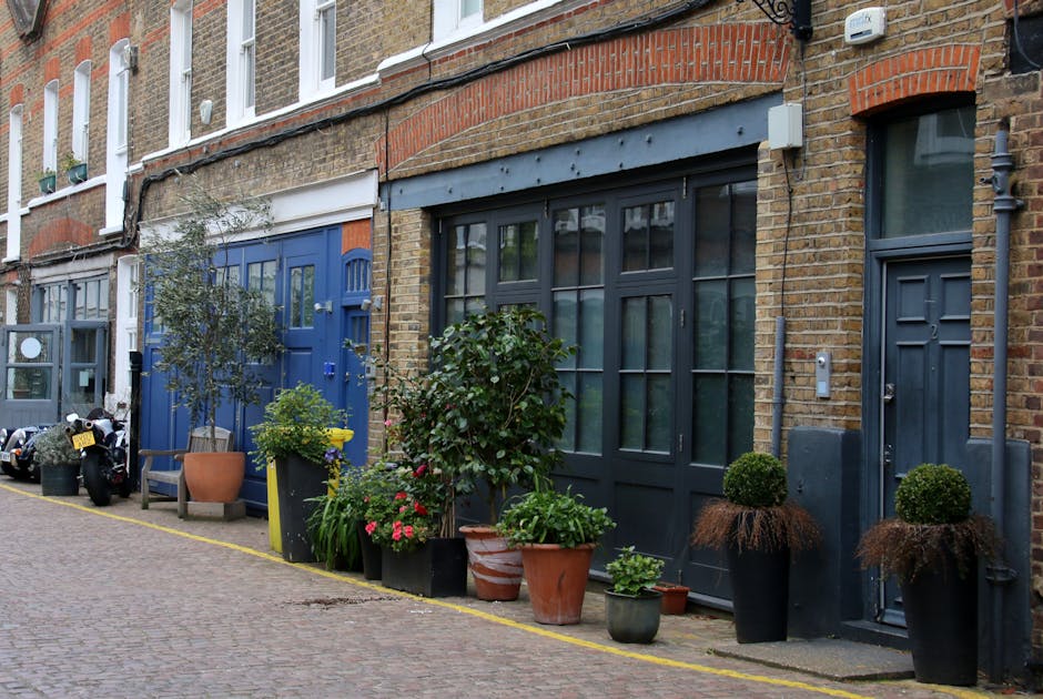 An urban garden along the side of a brick residential building on Kingsland Road, Dalston, featuring a diverse array of potted plants and shrubs arranged along the pavement. The plants include floweri